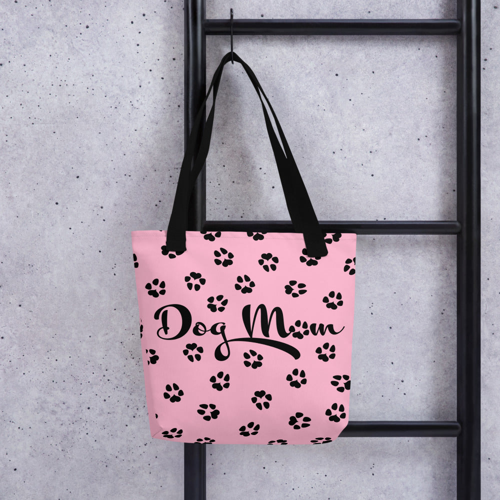 Tote bag with pink background. Dog Mom is written on the front with dog paw prints all over in black. The handle is black. The bag is shown hanging up.