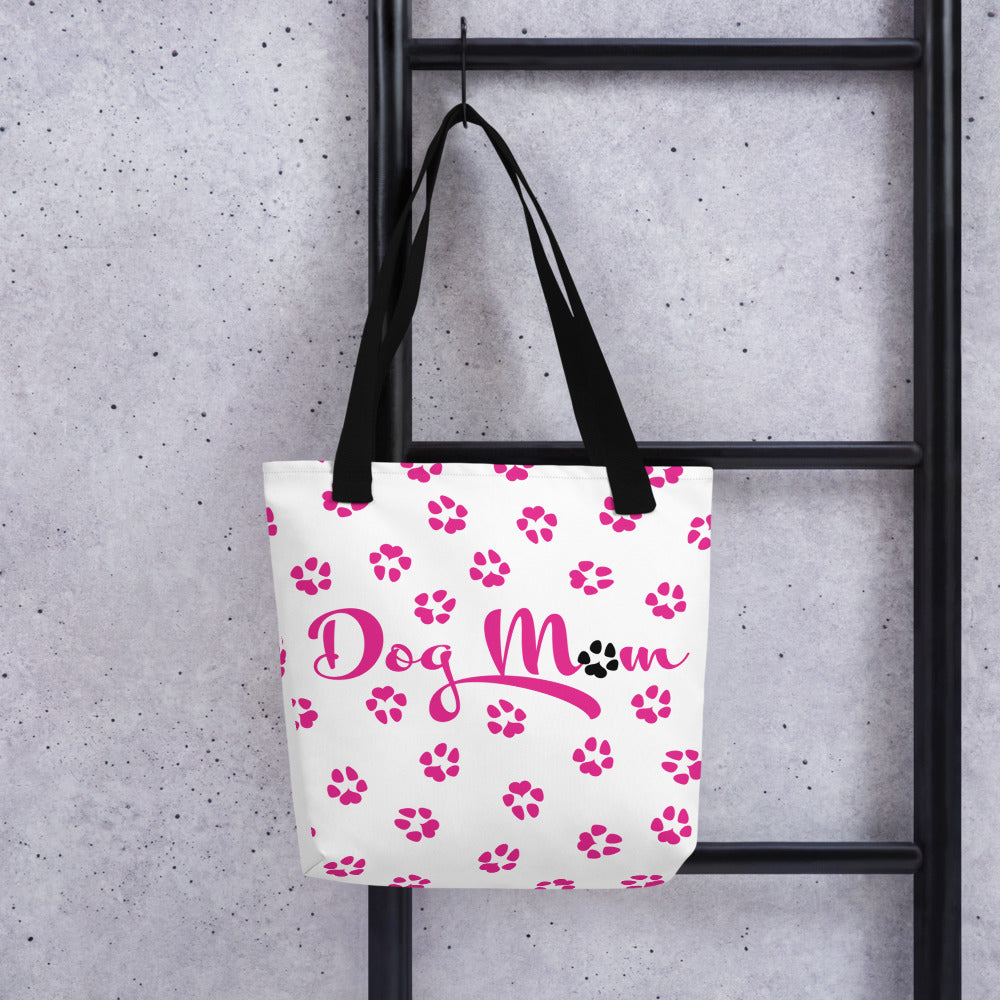 Tote bag with white background and black handle. Dog Mom is written on the front with dog paw prints all over in pink with one black paw in Mom. Bag is hanging up